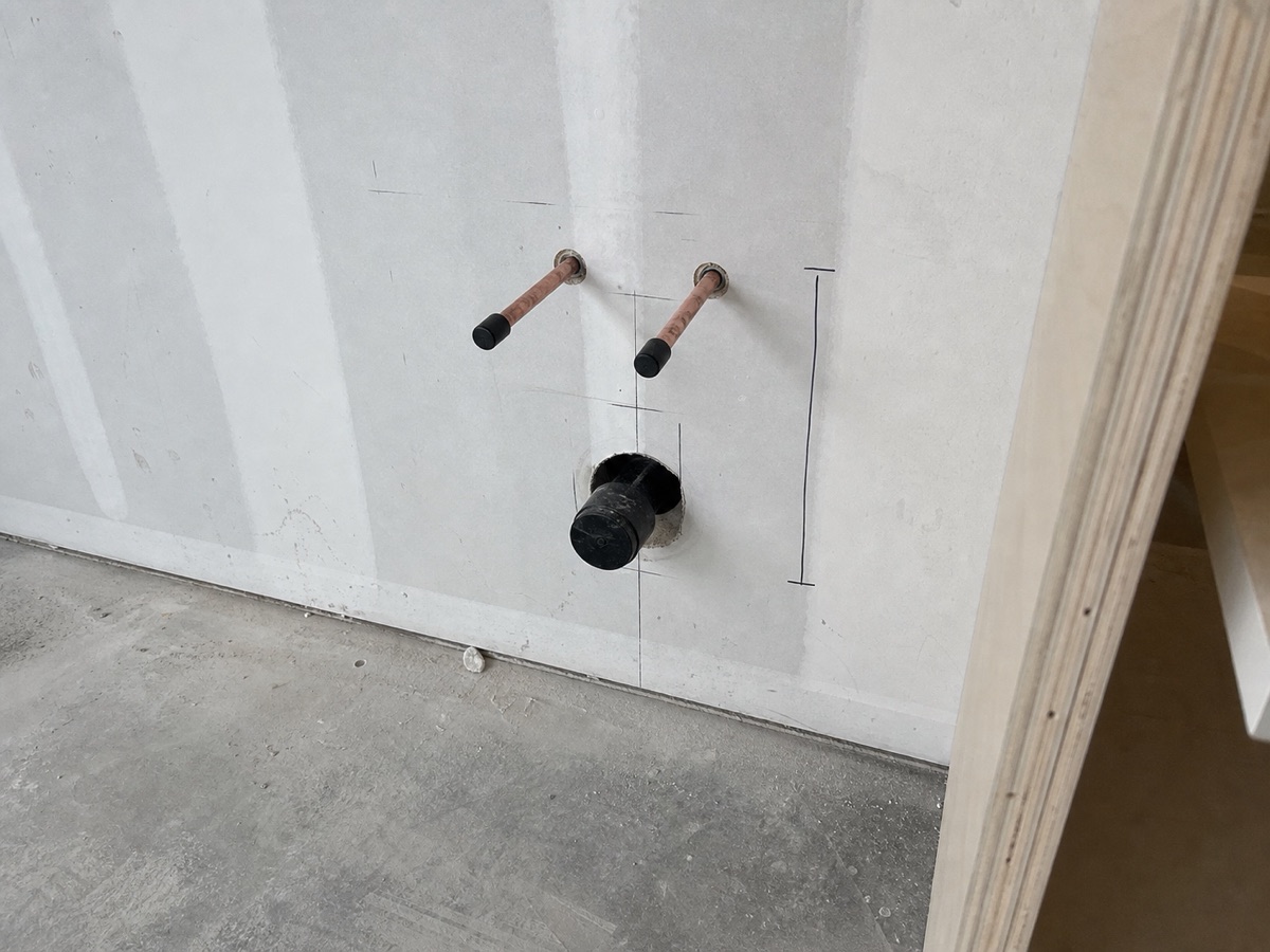 Site photo: two capped copper supply stubs and a drain rough-in on a boarded wall, with pencil layout marks